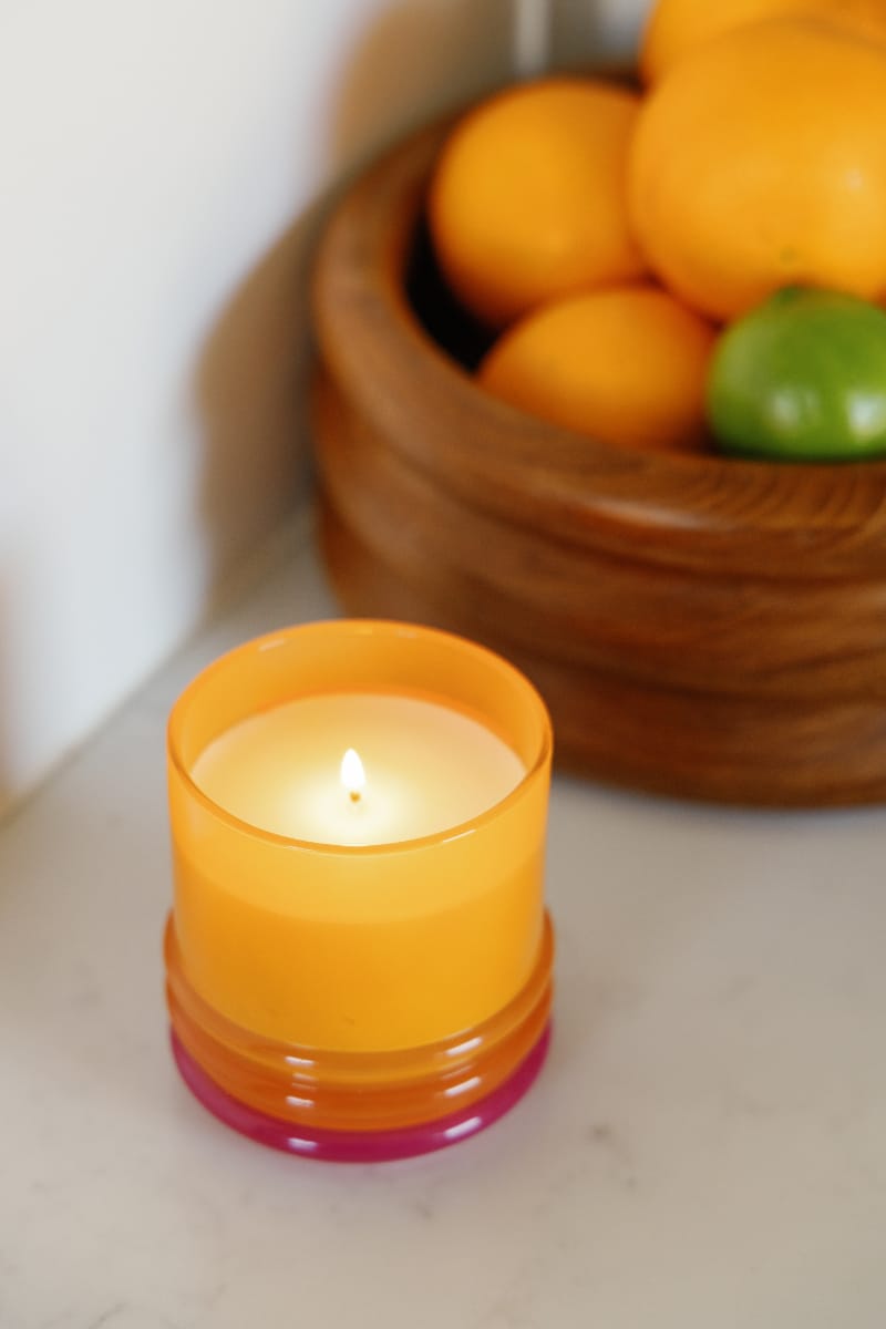 A lit Infrared Mango - Stack 7 oz Candle in an orange glass vessel sits on a white surface beside a wooden bowl filled with oranges and a green lime.