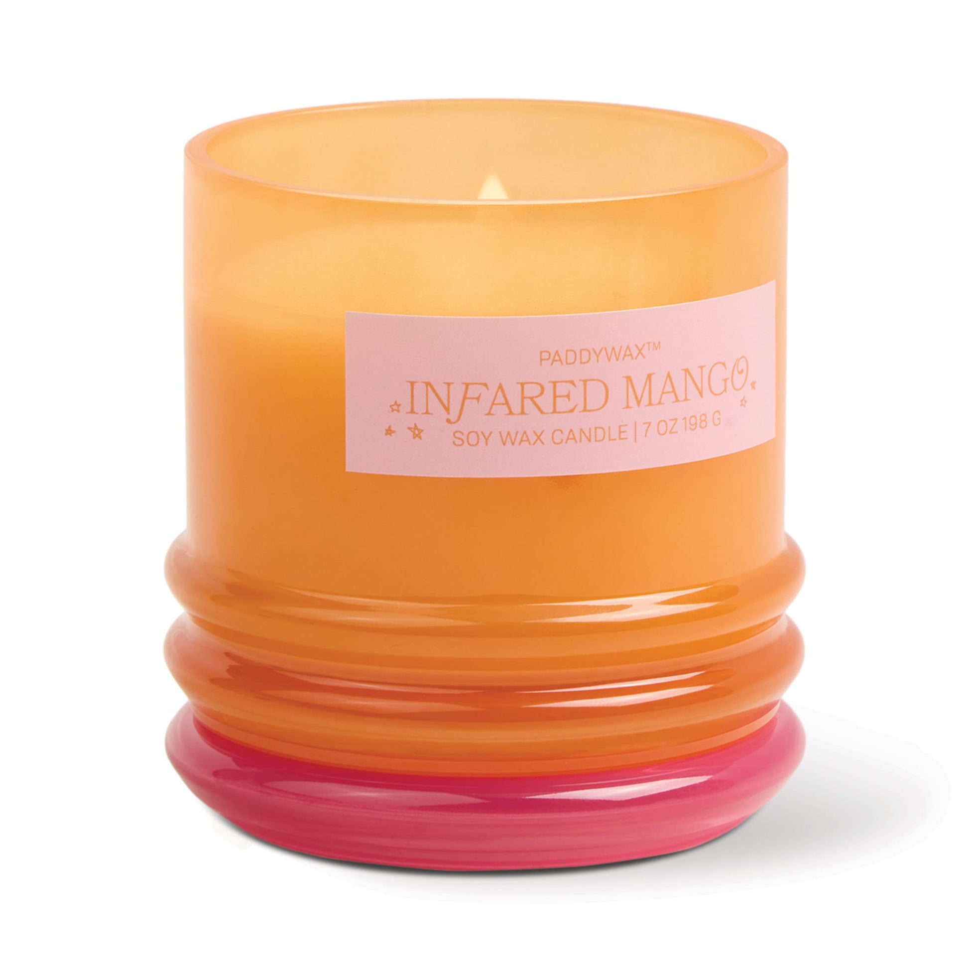 An orange glass vessel with a pink base holds the lit Infrared Mango - Stack 7 oz Candle. The label reads Paddywax, Soy Wax Candle, 7 oz/198 g. Once finished, repurpose the vessel for stylish storage.