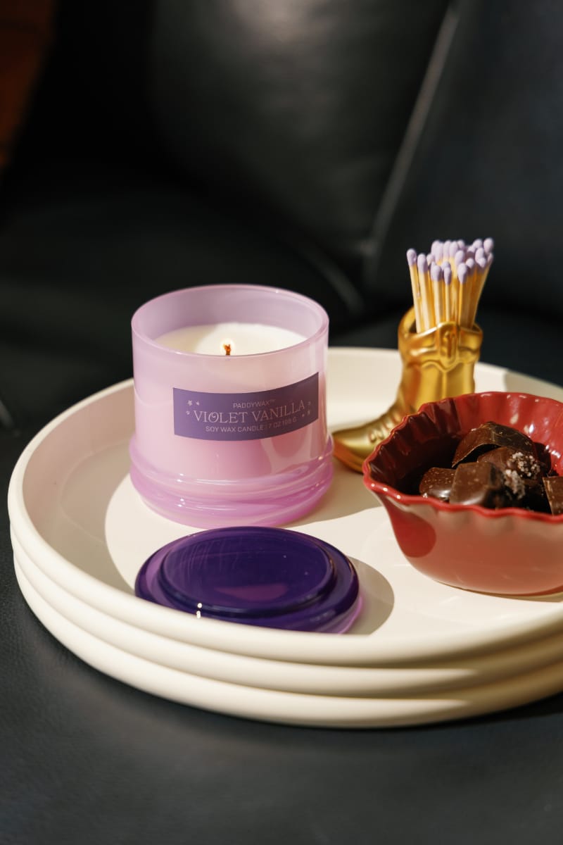 The Violet Vanilla - Stack 7 oz Candle, a pastel purple floral candle in a reusable glass vessel with a purple lid, sits on a white tray alongside a red bowl of chocolate treats and a gold match holder with matches on a dark surface.