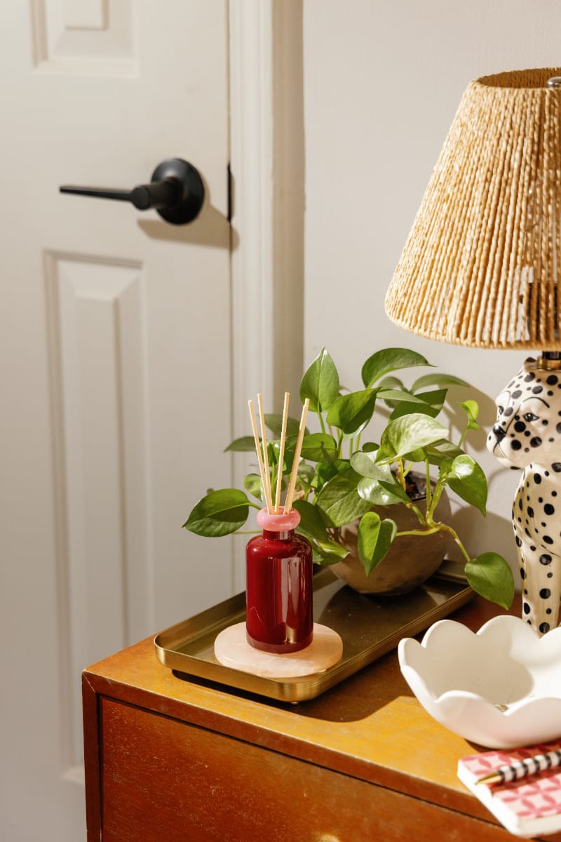 A modern bedside setup: the Saffron Rose - Stack 3.2 oz Diffuser for lasting fragrance, a mini potted plant, woven-shade lamp, and ceramic dish beside a white door with black handle—ideal for contemporary decor.