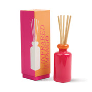 A pink and orange box labeled Infrared Mango - Stack 3.2 oz Diffuser stands beside a matching bottle with wooden reeds, filling the room with a sweet tropical fragrance.