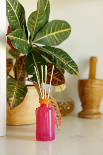 The Infrared Mango - Stack 3.2 oz Diffuser with an orange cap sits on a white surface by a leafy potted plant, emitting a tropical scent. In the background are a wooden mortar and pestle and a glass container.