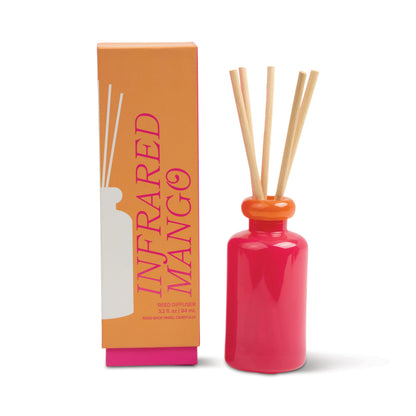 A bright pink Infrared Mango - Stack 3.2 oz Diffuser with natural reed sticks sits by its orange box labeled INFRARED MANGO in bold pink. The tropical scent includes vanilla bean and a minimalist white diffuser illustration decorates the packaging.