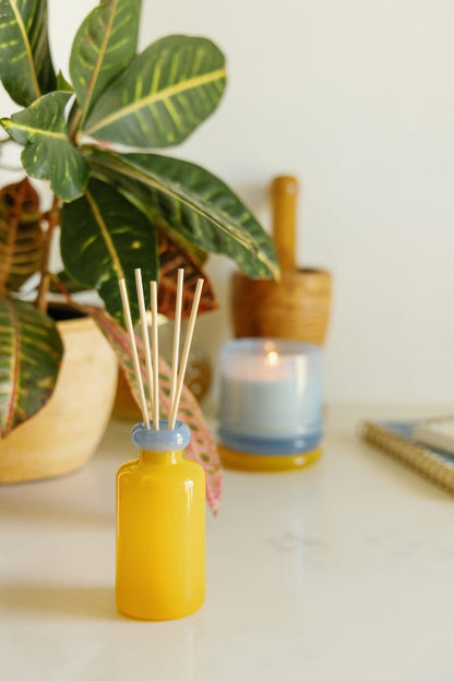 A Zesty Lemon - Stack 3.2 oz Diffuser with wooden sticks sits on a white surface, surrounded by a potted plant, a lit candle in a blue holder, and a wooden mortar and pestle for a modern home fragrance vibe.
