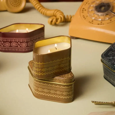 A Linen & Orris - Station Tin 3.5oz candle sits lit on a table among other tins, filling the air with its signature woody notes inspired by Northern California, while an old rotary phone and coiled cord lend vintage charm in the background.
