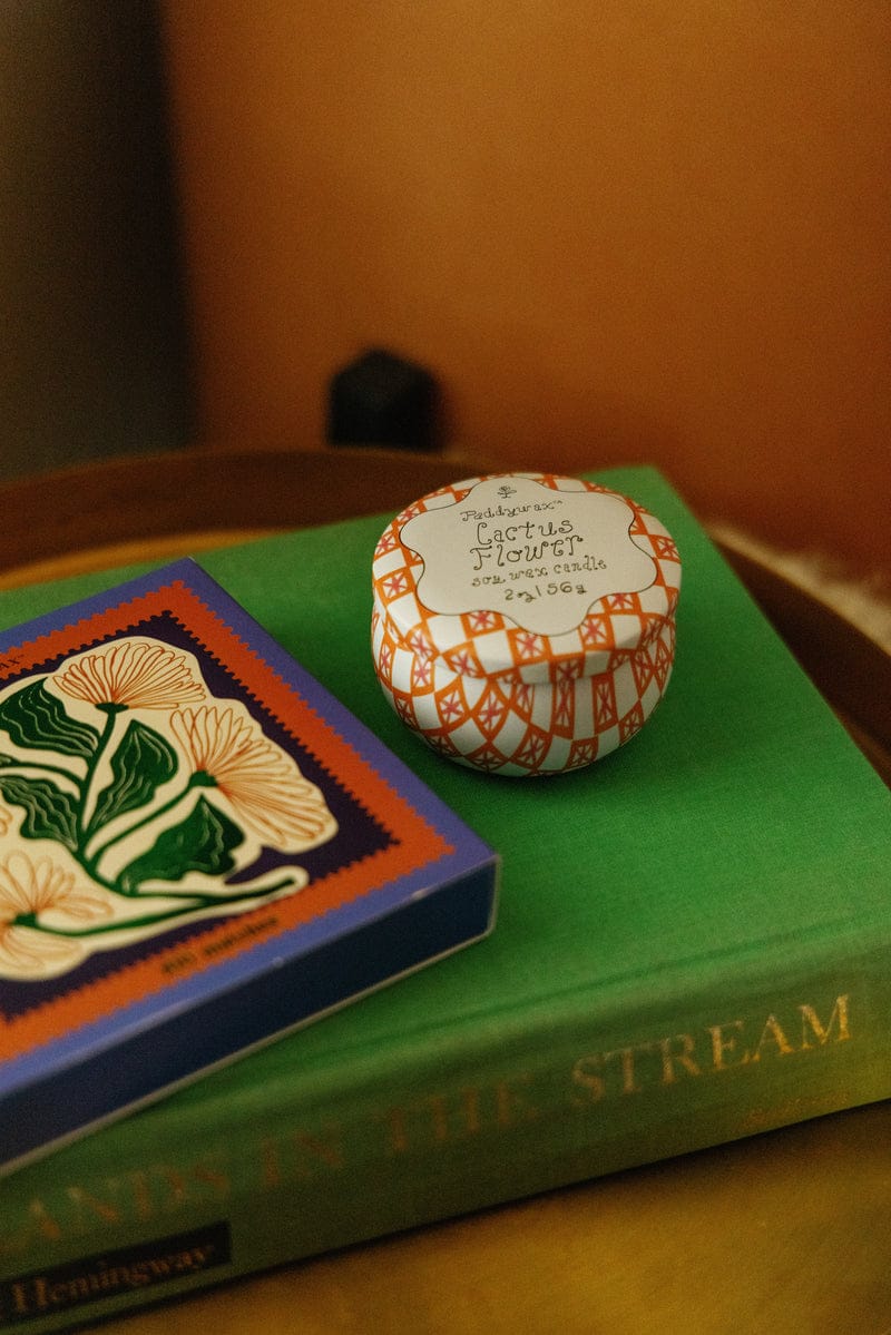 A Cactus Flower - Tulip 2 oz Tin Candle, with luminous notes of cactus flower and desert blooms, sits on a green book with gold lettering next to a colorful book featuring a botanical illustration.
