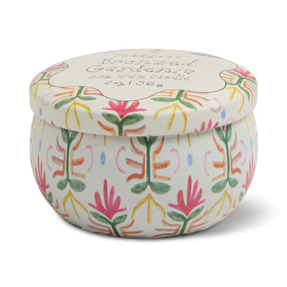 The Bronzed Gardenia - Tulip 2 oz Tin Candle features a round, white tin adorned with pink, green, and yellow floral patterns. It evokes creamy florals and sunlit citrus, with a matching lid topped by cursive writing.
