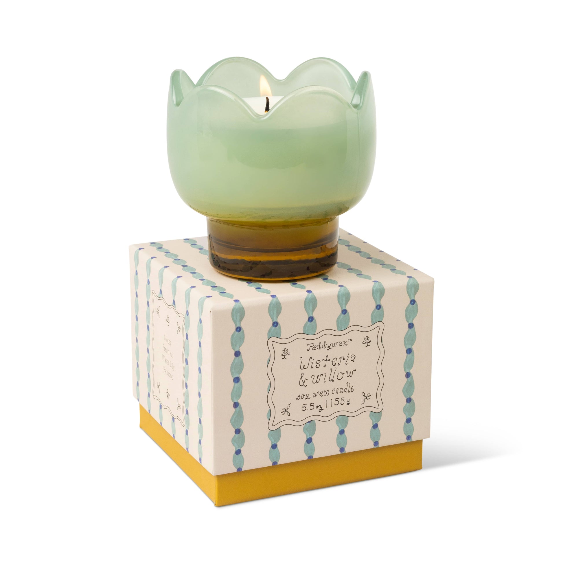 A lit Wisteria & Willow - Tulip 5.5 oz soy wax candle in green scalloped glass sits atop a cream and yellow box with blue bead detailing. The decorative box is labeled "Lusterie & Willow, soy wax candle, 5.5 oz.