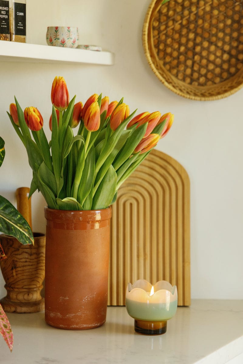 A terracotta vase with red and yellow tulips sits on a white countertop beside a lit Wisteria & Willow - Tulip 5.5 oz Candle in a decorative holder, with a woven tray and arched wooden panel in the background.