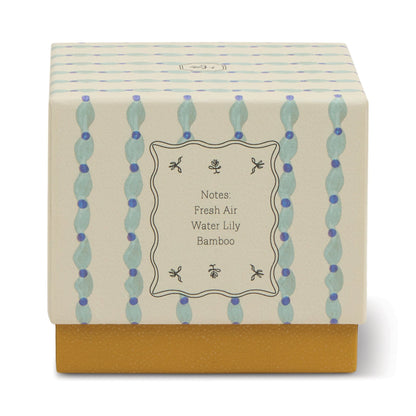 The Wisteria & Willow - Tulip 5.5 oz Candle comes in a decorative botanical box with a cream and blue patterned lid labeled Fresh Air, Water Lily, Bamboo. Its solid mustard yellow base is perfect for teal glass vessels or wisteria candles.