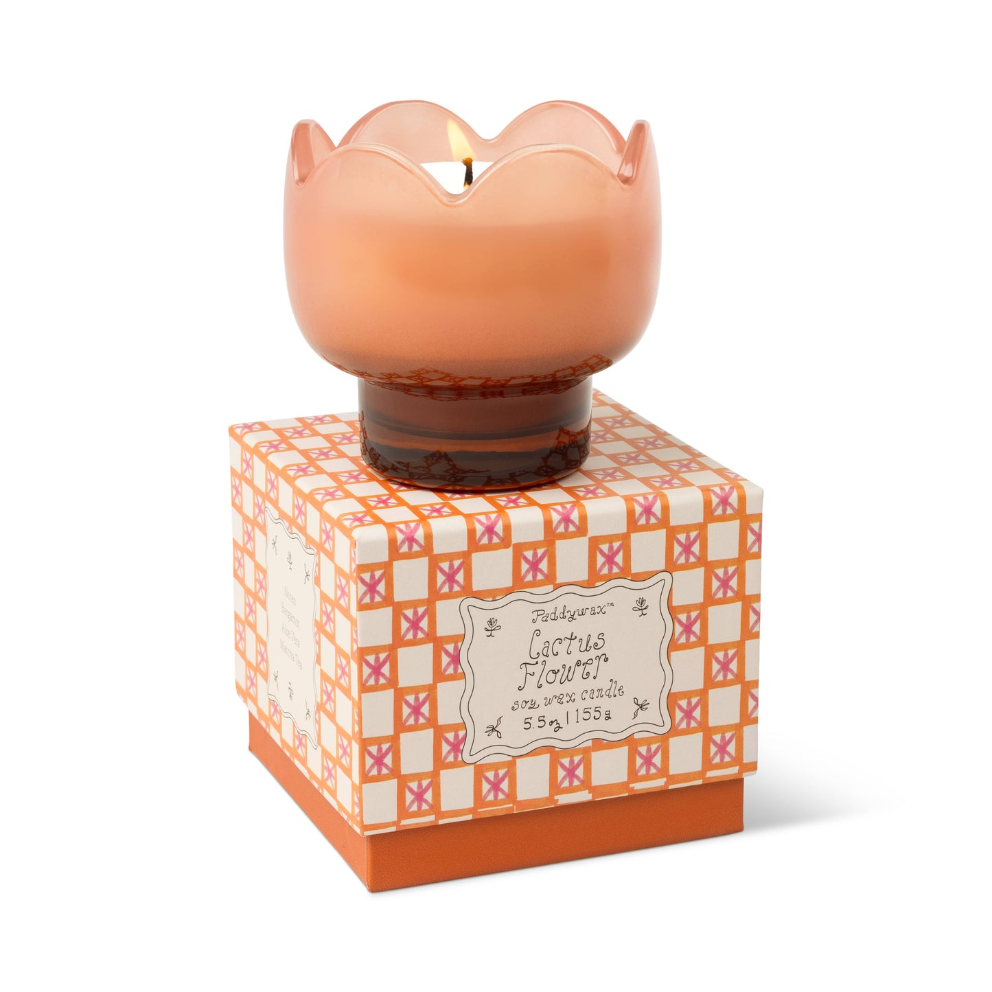 A lit Cactus Flower - Tulip 5.5 oz Candle in a pink, scalloped glass holder rests on a patterned orange and white box with desert bloom-inspired designs, making it a perfect decorative accent with detailed product info included.