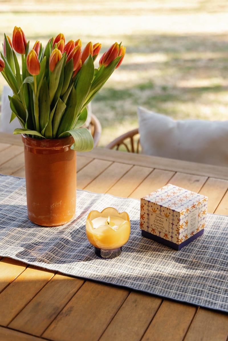 A brown rustic vase with orange tulips, the Garden & Grove - Tulip 5.5 oz Candle, and a decorative box sit on a checkered table runner atop an outdoor wooden table with blurred greenery in the background.