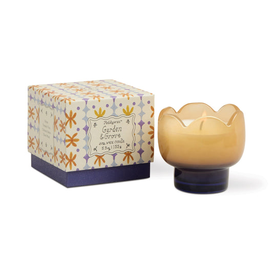 The Garden & Grove - Tulip 5.5 oz Candle sits in a rustic vessel beside its purple floral-patterned box, featuring orange and lavender flowers—perfect for garden-inspired home decor.