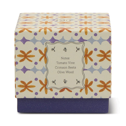 The Garden & Grove - Tulip 5.5 oz Candle comes in a decorative square box with floral and geometric patterns in orange, purple, and gray. Enjoy garden-inspired aromas of tomato vine, crimson beets, and olive wood in a rustic vessel.