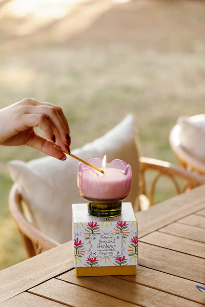 A hand lights the Bronzed Gardenia - Tulip 5.5 oz Candle, shaped like a pink flower, atop a decorative floral box on a wooden outdoor table with chairs and cushions—ideal for adding floral charm to your home decor.