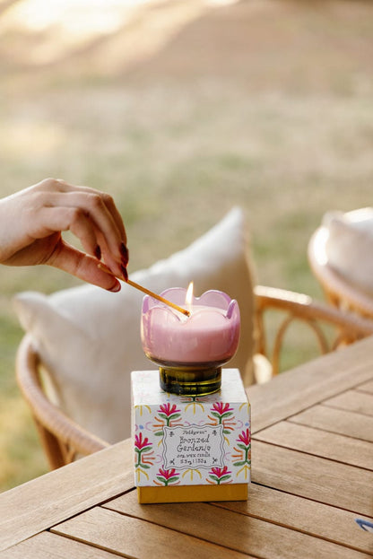 A hand lights the Bronzed Gardenia - Tulip 5.5 oz Candle, shaped like a pink flower, atop a decorative floral box on a wooden outdoor table with chairs and cushions—ideal for adding floral charm to your home decor.