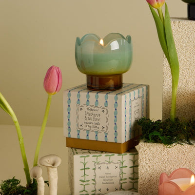 The Wisteria & Willow - Tulip 2 oz Tin Candle sits on patterned boxes, surrounded by pink tulips, moss, ceramic mushrooms, and other candles. Its pastel tin and spring scent create a charming display against a soft beige background.