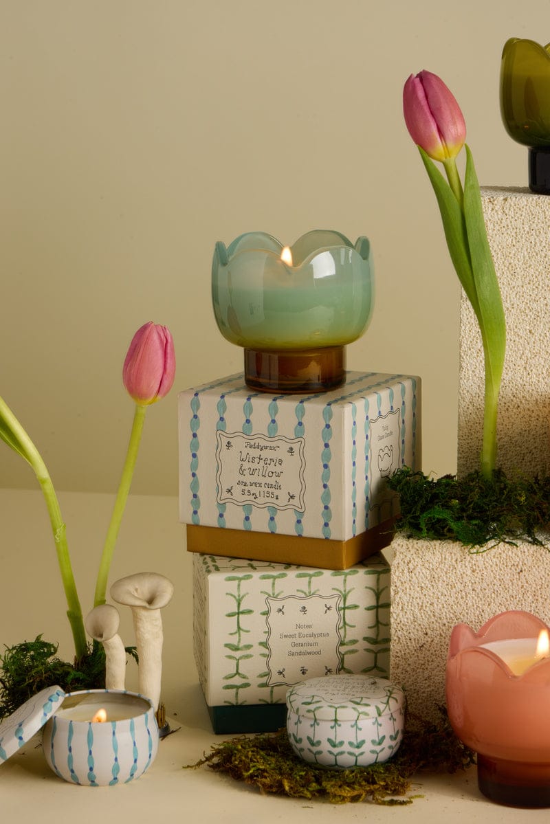The Wisteria & Willow - Tulip 2 oz Tin Candle sits on patterned boxes, surrounded by pink tulips, moss, ceramic mushrooms, and other candles. Its pastel tin and spring scent create a charming display against a soft beige background.