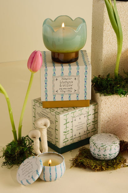 A decorative candle in a green glass holder sits on a patterned box with moss, a pink tulip, and small mushrooms. Nearby is the Verbena & Lemongrass - Tulip 2 oz Tin Candle and stacked boxes.