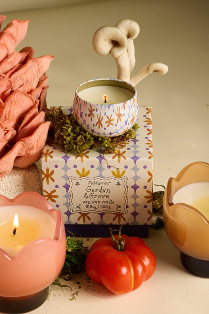 The Garden & Grove - Tulip 2 oz Tin Candle rests on a floral box amid moss, a tomato vine, a pink flower, and two glass candles, with two mushrooms growing behind the patterned tin cup.