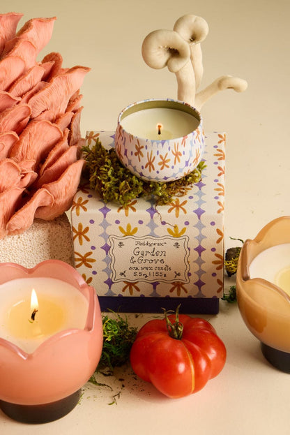 The Bronzed Gardenia - Tulip 2 oz Tin Candle, in a patterned holder atop a Garden & Grove box, is surrounded by two lit candles, moss, mushrooms, and a ripe tomato, filling the air with creamy florals and sunlit citrus.