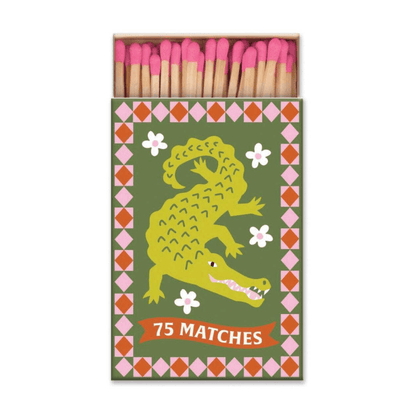 The A Dopo "Crocodile" - 75ct Boxed Matches features a crocodile illustration, floral accents, and a “75 Matches” banner. The open box reveals pink-tipped matches—ideal for lighting your favorite A Dopo candle.