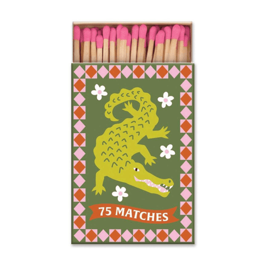 The A Dopo "Crocodile" - 75ct Boxed Matches features a crocodile illustration, floral accents, and a “75 Matches” banner. The open box reveals pink-tipped matches—ideal for lighting your favorite A Dopo candle.