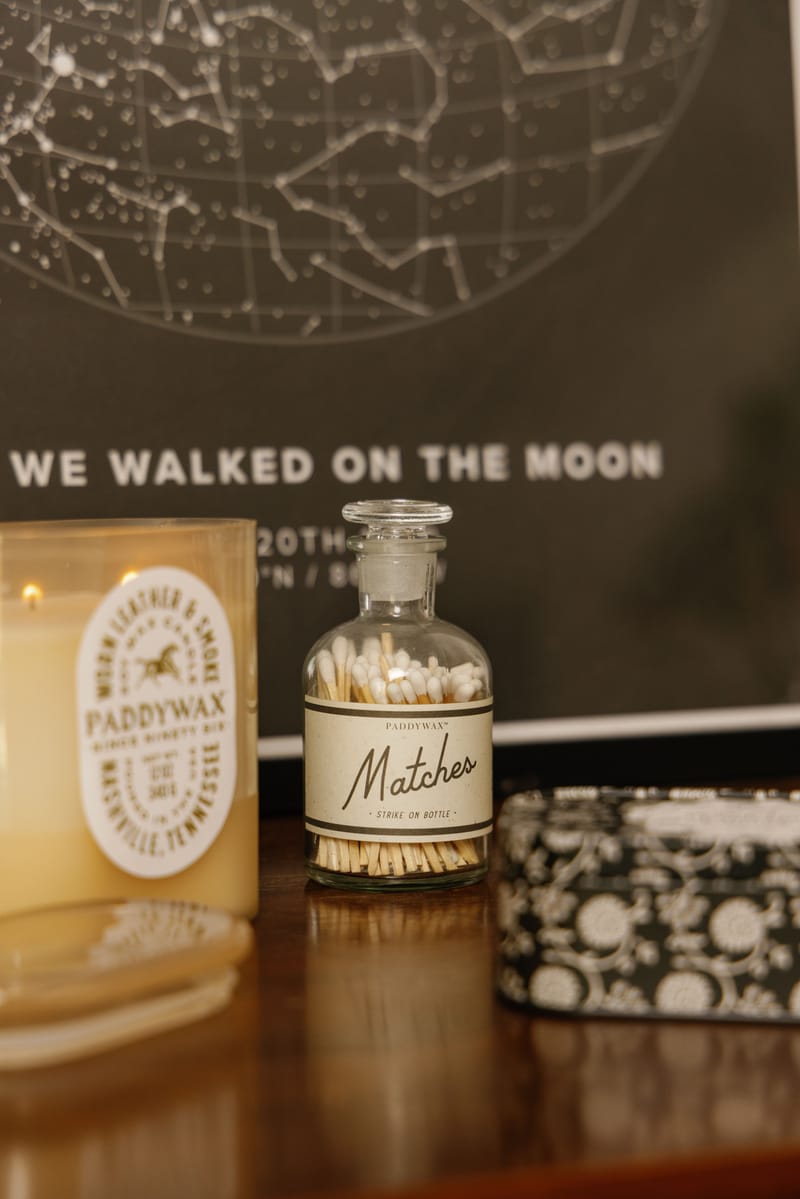 A close-up of a shelf features a Bottle of Matches filled with wooden matches, a candle, a decorative box, and part of a star map print in the background with the words WE WALKED ON THE MOON.