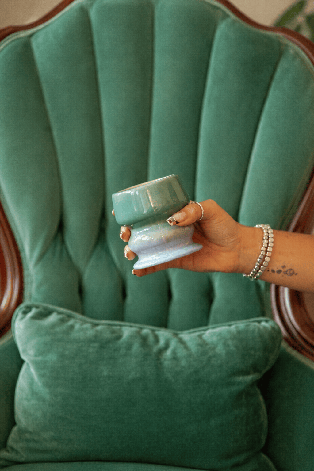 A hand holding a green ceramic cup with a white base, in front of a green tufted armchair with a matching pillow. The hand, adorned with bracelets and a small tattoo, captures the serene atmosphere created by the Fresh Air & Sea Salt - Melt 8.5 oz Candle, evoking the subtle coastal fragrance drifting through the room.