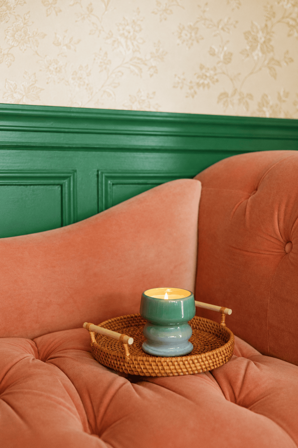 Perched on a wicker tray atop a plush, peach-colored sofa, the Fresh Air & Sea Salt - Melt 8.5 oz Candle in its green ceramic holder infuses the room with coastal fragrance, while green paneling and cream floral wallpaper complete the serene setting.