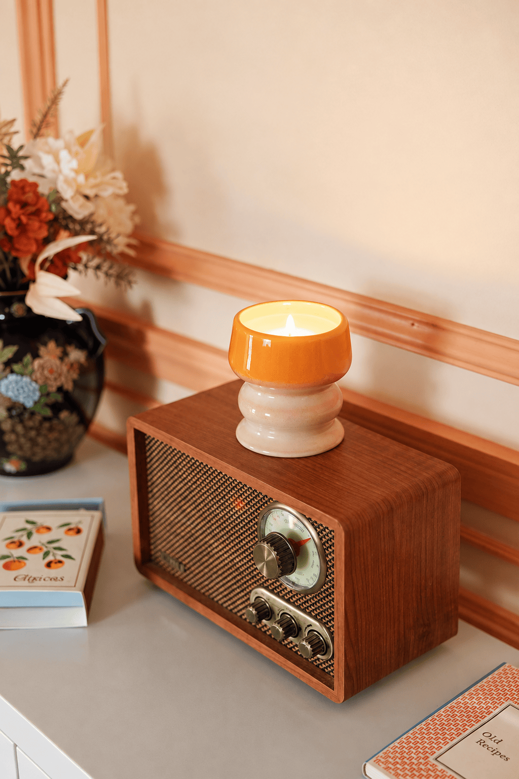 A vintage radio sits on a table beside a hand-glazed ceramic vessel and two closed notebooks with decorative covers, while the air is filled with the tropical scent of the lit Infrared Mango - Melt 8.5 oz Candle.