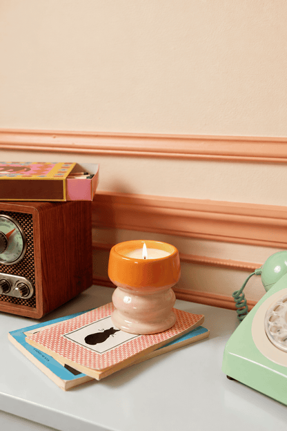 An Infrared Mango - Melt 8.5 oz Candle in a hand-glazed ceramic vessel burns atop a notebook near a retro radio, a stack of books, and a vintage green rotary phone on a white surface set against beige and peach walls, infusing the space with its vibrant tropical fragrance.