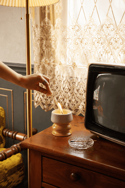 A hand lights the Smoked Pistachio - Melt 8.5 oz Candle on a wooden table next to an ashtray and an old television, with lace curtains and a vintage lamp in the background.