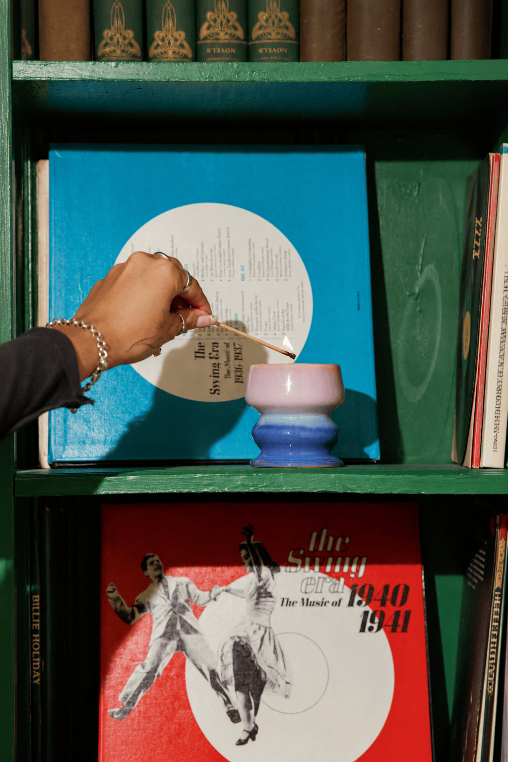 A hand holding an incense stick over a purple and blue holder on a bookshelf, with vinyl records, books, and the repurposable vessel from a Teakwood & Jasmine - Melt 8.5 oz Candle—now uniquely displayed—gracefully arranged in the background.
