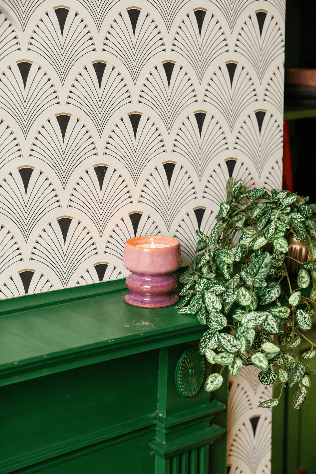 A glowing Wild Fig & Cedar - Melt 8.5 oz Candle in a pink holder rests on a green mantel beside a leafy potted plant, with art deco-style wallpaper providing the backdrop.