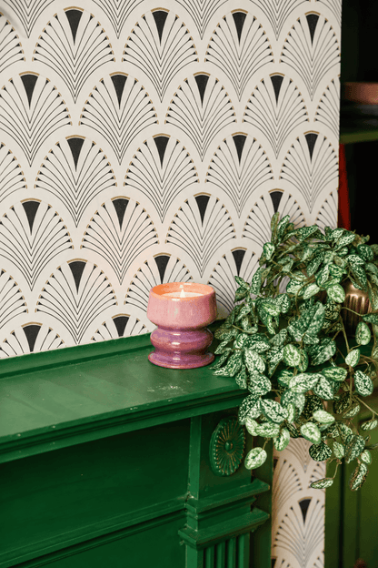A glowing Wild Fig & Cedar - Melt 8.5 oz Candle in a pink holder rests on a green mantel beside a leafy potted plant, with art deco-style wallpaper providing the backdrop.