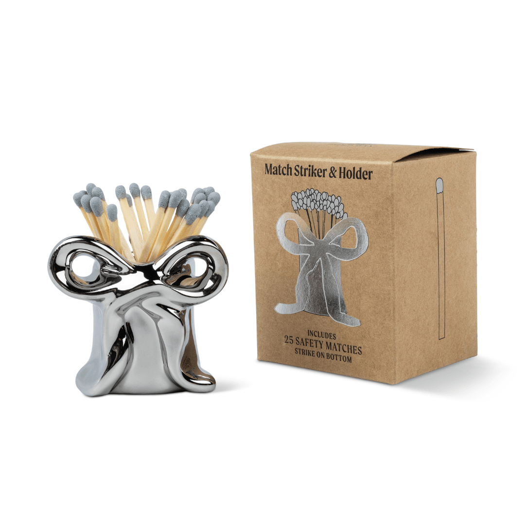 A Metallic Bow Match Holder, crafted in shiny silver and filled with safety matches, sits beside its brown box packaging labeled “Match Striker & Holder” and featuring a match illustration.