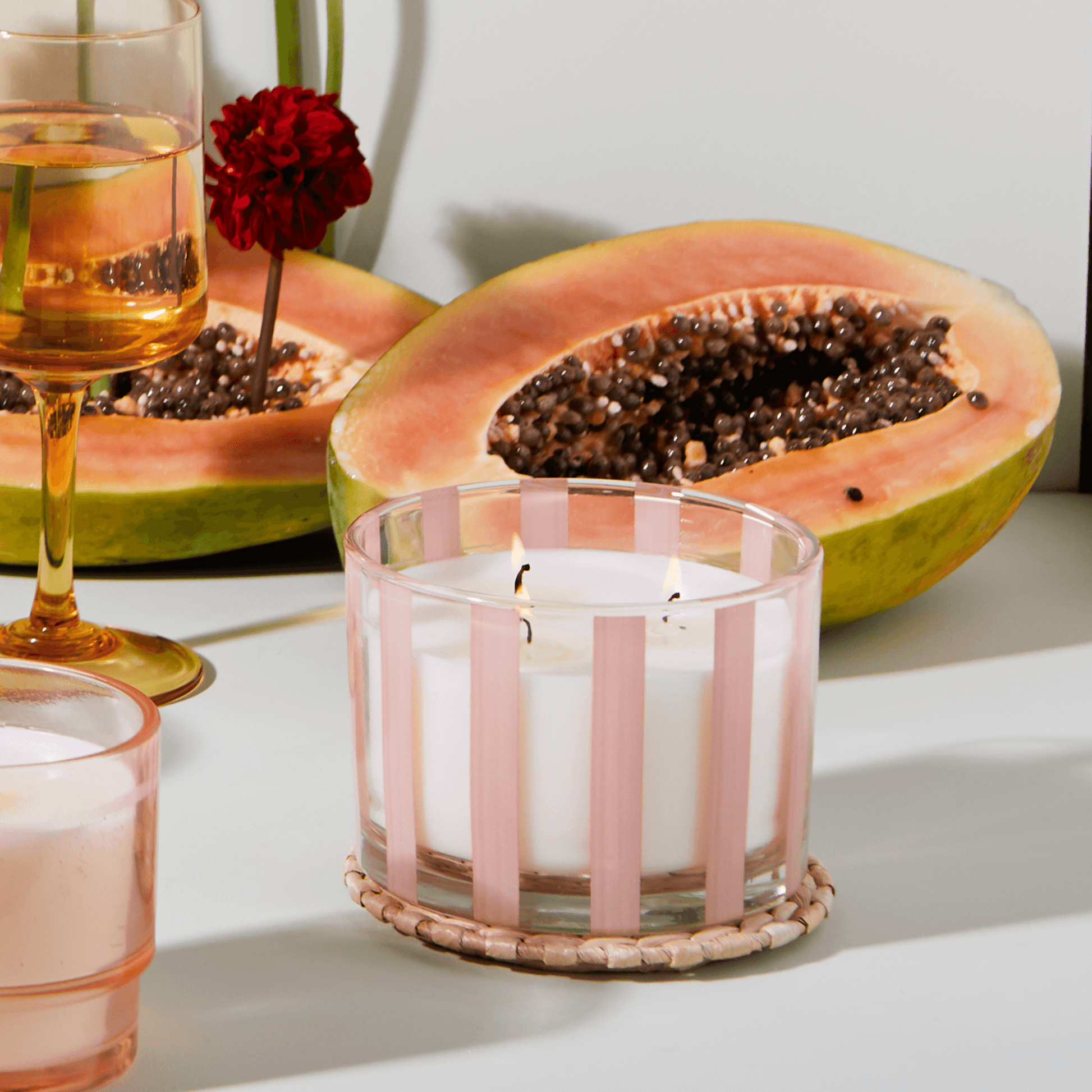 12 oz clear glass vessel with light pink stripes, white wax, and three wicks; a part of the Al Fresco collection; comes with woven rattan lid; pictured lit in front of a papaya