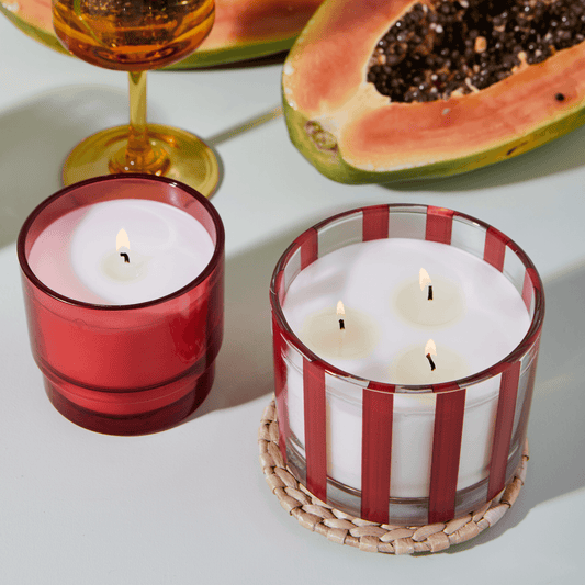 a12 oz clear glass vessel with red stripes, white wax, and three wicks; a part of the Al Fresco collection; comes with woven rattan lid; pictured sitting on the rattan lid, lit next to a smaller, solid red candle of the same color