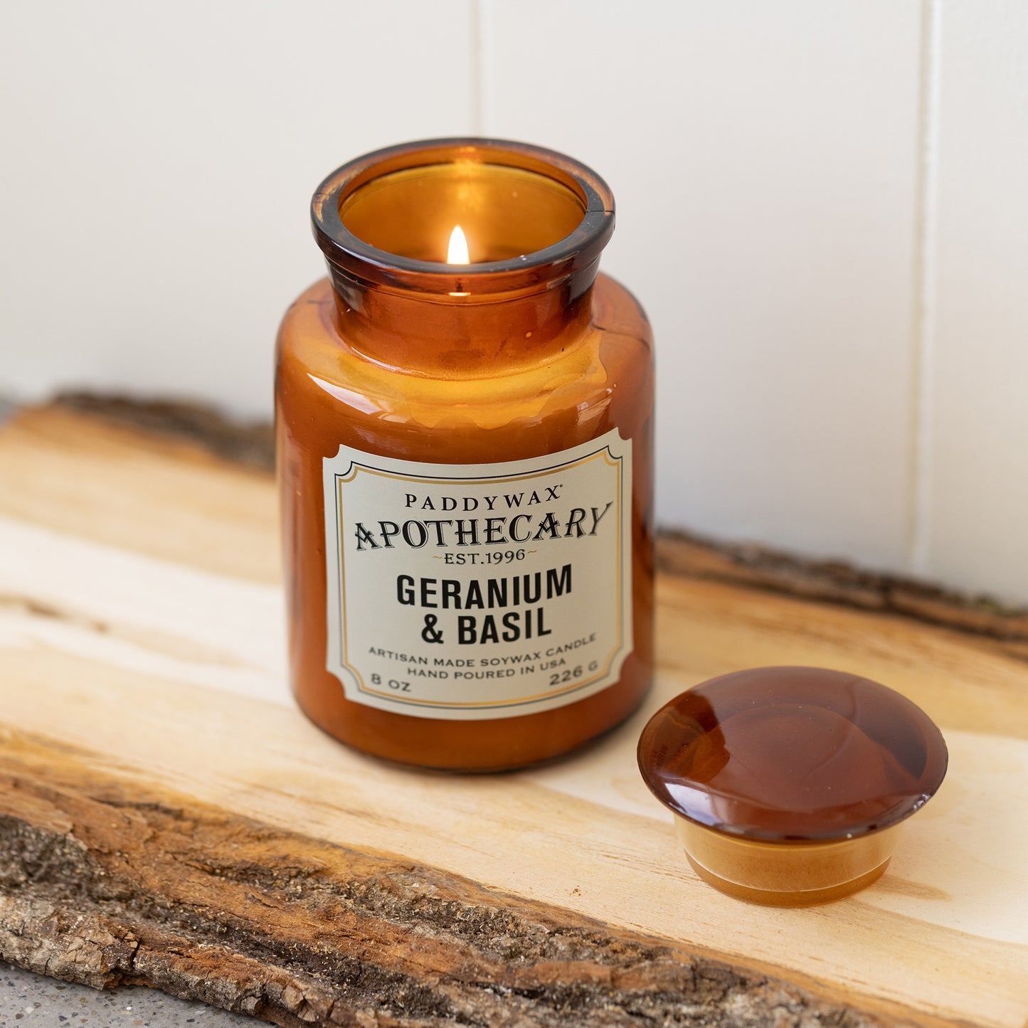 8 oz brown apothecary bottle glass vessel with a vintage-style label; white wax; part of the apothecary collection; candle lid off and scented candle burning on wood piece