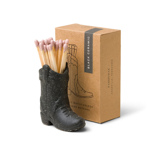 small black ceramic cowboy boot pictured holding pink-tipped matches in front of the brown packaging