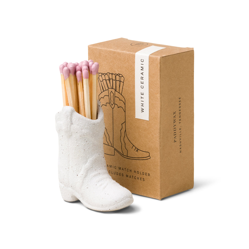 small white ceramic cowboy boot pictured holding pink-tipped matches in front of the brown packaging