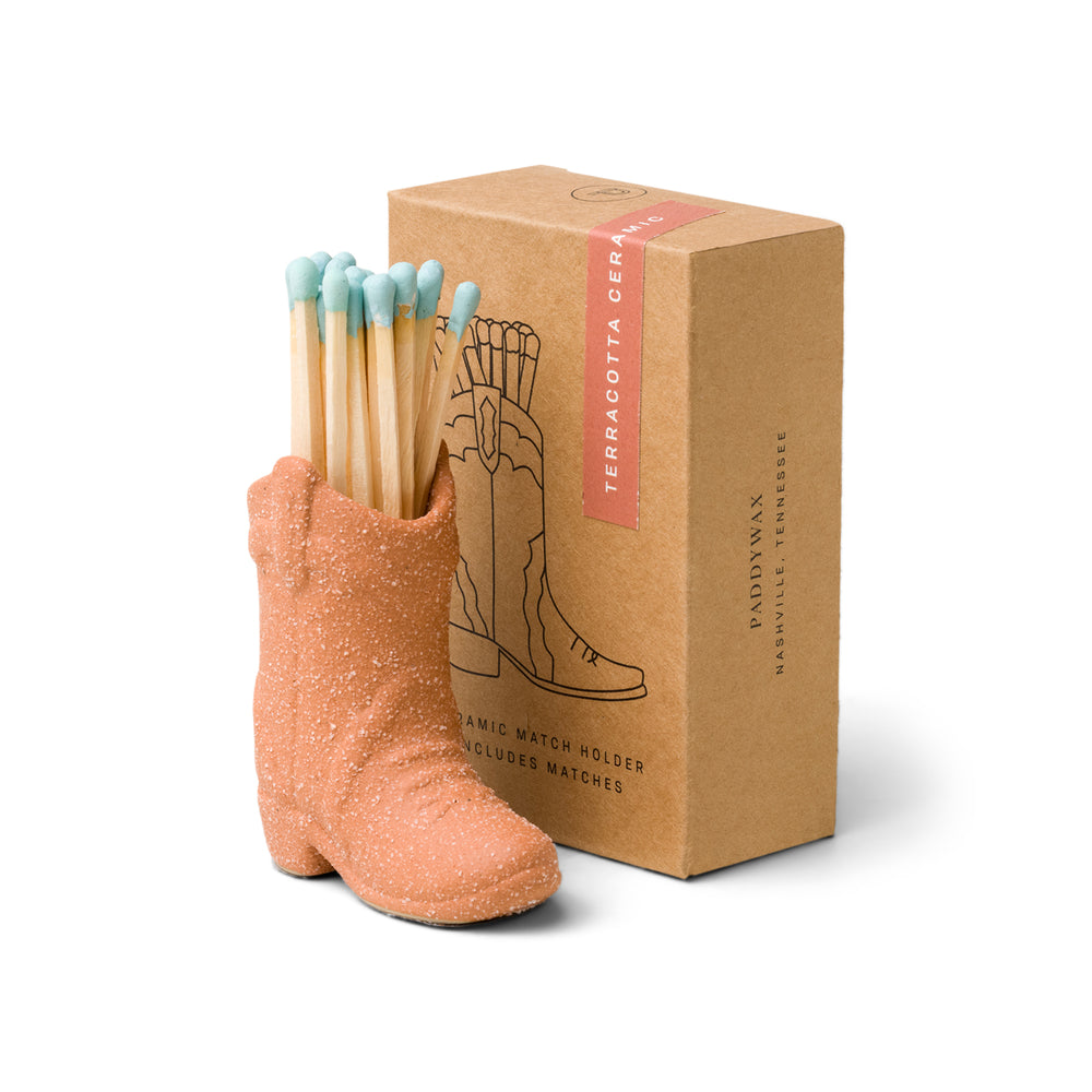 terracotta (desert orange) ceramic cowboy boot pictured holding blue-tipped matches in front of the brown packaging