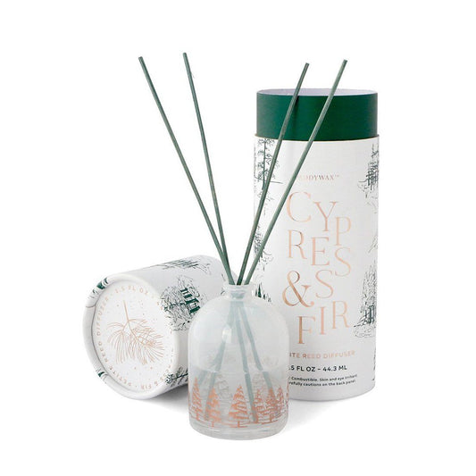 hazy white diffuser bottle with copper colored fir trees, holding sticks; pictured next to cylindrical white gift box for candle with green fir trees