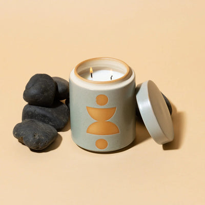 The Ocean Rose + Bay - Form 12 oz Candle, in a ceramic planter with an abstract yellow design and matching lid, glows beside smooth black stones on a beige surface, adding warmth and fragrance to any space.