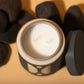 12 oz black cylindrical ceramic vessel with ceramic lid; white wax and two cotton wicks; reflected beige semi-circle design on the side; pictured with lid off