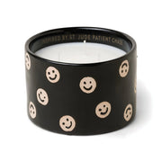 The Black Fig + Olive - St. Jude Giveback "Celebrate" 11 oz. Candle features a black ceramic holder with beige smiley faces, white wax, and INSPIRED BY ST JUDE PATIENT CHASE inscribed inside, making it both meaningful and unique.
