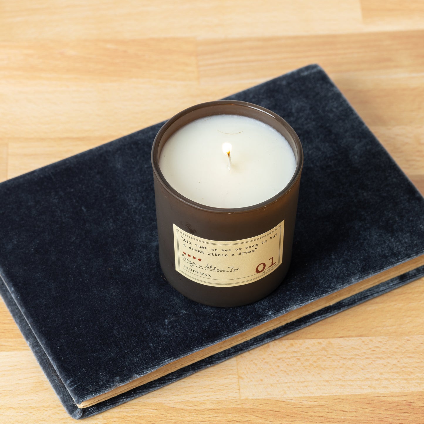 Overhead shot of Library Edgar Allan Poe soy wax candle with single wick burning on top of book on hardwood surface