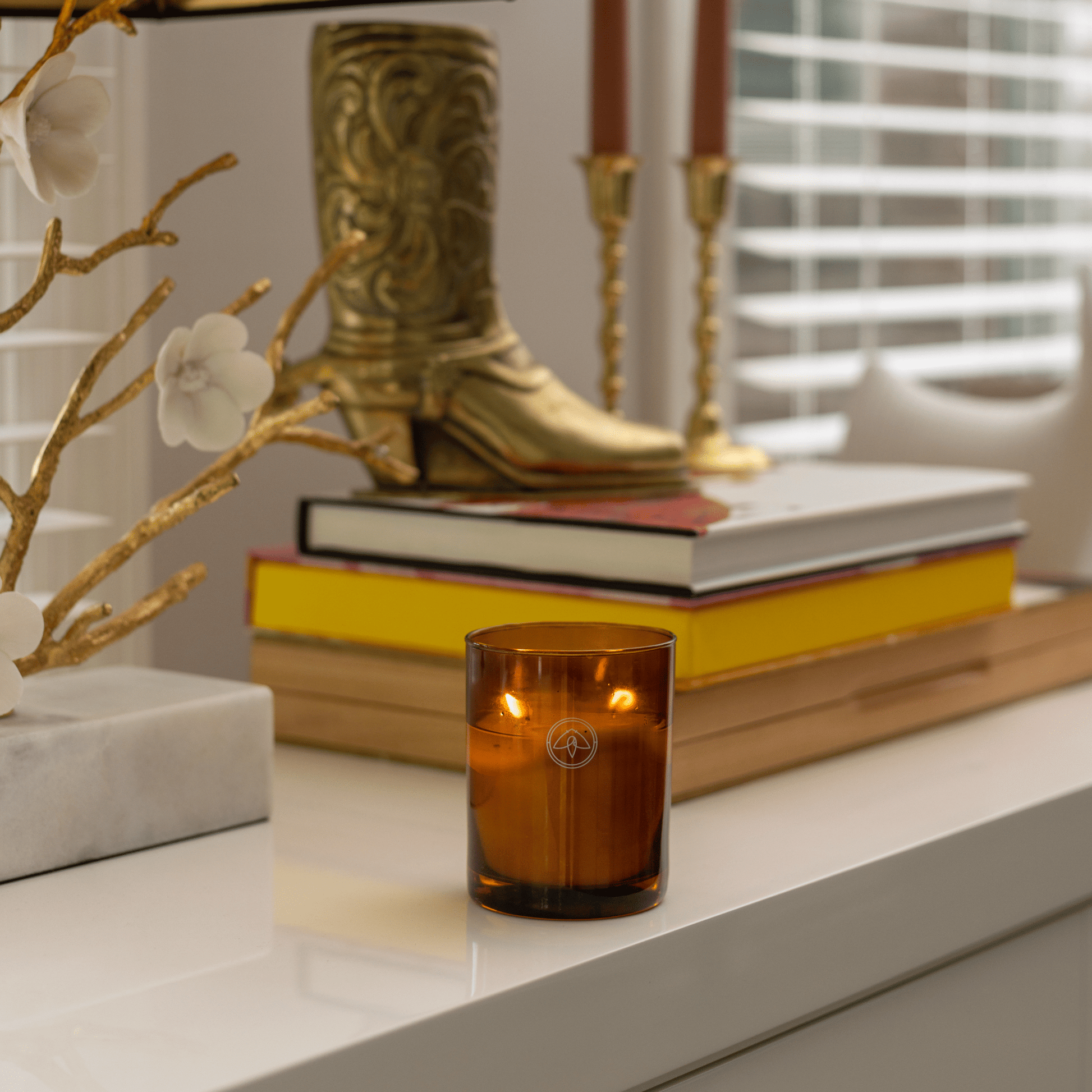 10oz Glow two wick canlde on white table with books, metal boot decor and candlesticks with window in background