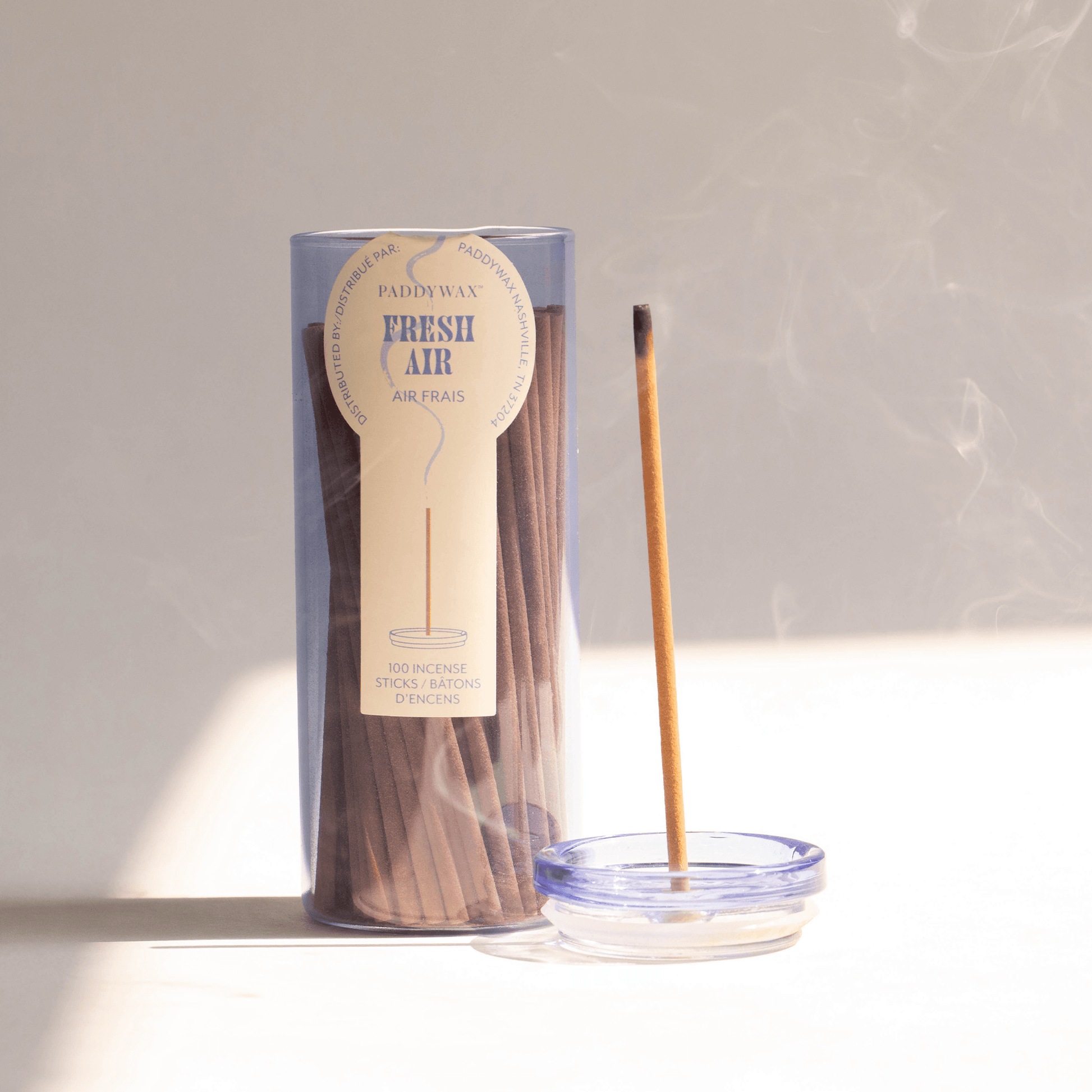 Blue glass cylinder with white label holding 100 incense sticks for flameless fragrance; pictured next to burning incense stick